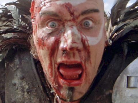 A man covered in blood and cuts is looking straight at the camera and shouting. He is wearing amour and feathers around his neck.