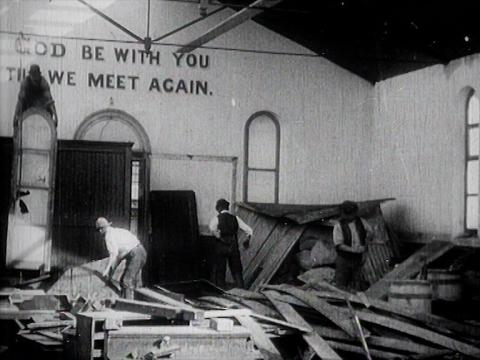 Three workers are demolishing the interior of a church. On one of the walls, which is still standing, it is written 'God Be With You Till We Meet Again'.