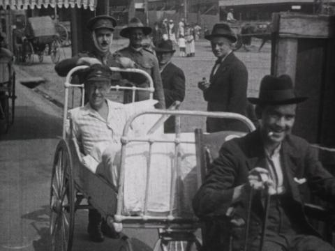 An injured soldier from the First World War is being wheeled on a trolley bed to a special cinema screening. He is surrounded by men, some in military uniform, looking at the camera.