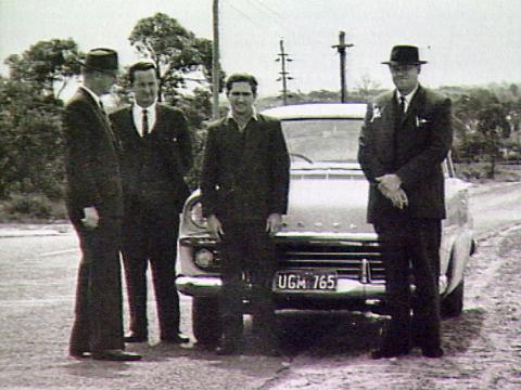 Three police detectives standing with serial killer Eric Edgar Cooke in front of a car on the side of the road. 