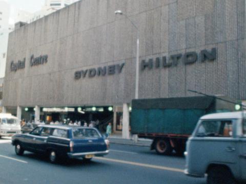 Front facade of the Sydney Hilton Hotel George Street entrance in 1978. It is a large concrete structure with 'SYDNEY HILTON' signage on the front. Some cars and trucks can be seen on the street in the foreground. 
