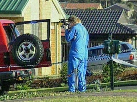 Two police forensic officers stand in the driveway of a house taking photographs behind a 4-wheel drive vehicle. The area photographed is surrounded by police tape. 