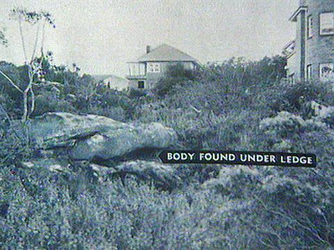 An area of scrubland with bushes and trees and a large rock in the centre. There is a text overlay on the image pointing to the rock that says 'Body found under ledge'. There are some houses in the background. 