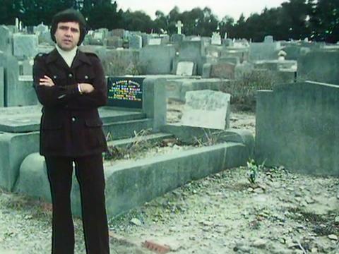 Full length shot of journalist Peter Luck standing with arms folded looking directly at camera in a cemetery with graves and headstones behind him. 