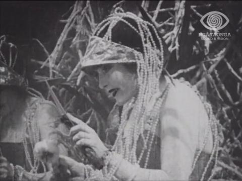 Annette Kellerman is looking at herself in a handheld mirror. She is dressed in a pearl headdress.