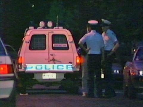 The rear of a police van park in the middle of a street. Two uniformed police officers stand to the right rear of the van. 