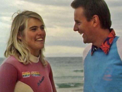 Australian surfer Pam Burridge wearing a pink wetsuit and Wonder World reporter Phillip Tanner, pictured from chest up standing on a beach with the ocean in the background.