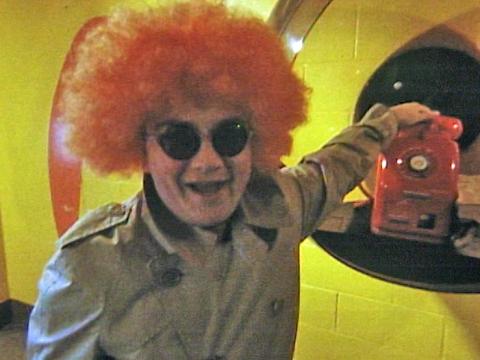 Jonathan Coleman wearing an orange fuzzy wig and a trenchcoat and sunglasses filming a report for Simon Townsend's Wonder World.