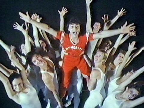 A group of dancers wearing white leotards with arms outstretched. In the centre is singer John St Peeters, in a red jumpsuit being lifted up by some of the dancers.