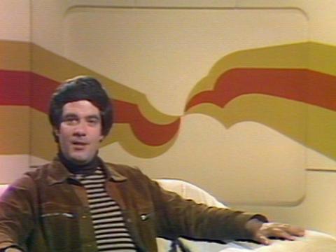TV presenter Simon Townsend, seated on a couch and looking at camera in a TV studio.