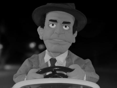 Black and white still of a male puppet in a suit and hat behind a steering wheel.