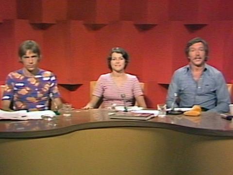 Three television presenters (two men and a woman) sit at a desk in a television studio facing directly towards the camera.