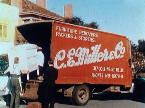 Two men are moving a piece of furniture into a big red removalist van with the name C.E. Miller & Co written on the side of it. Circa 1946.