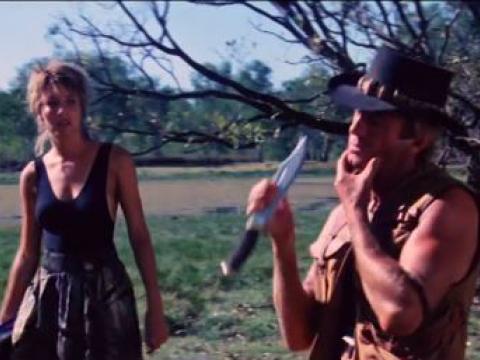 A rough looking man shaves with a large knife while a woman walks past looking shocked