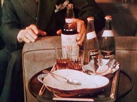 A side table next to the arm of a sofa. On the table are some glasses, ice, a bottle of Pimms and some soda water. You can see a man in a business suit sitting on the sofa about to pick up the bottle.