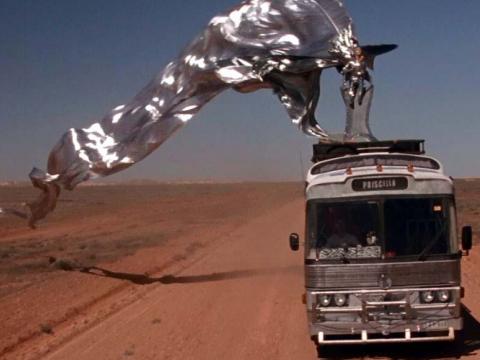 Guy Pearce with a long flowing silver banner riding on top of a bus in the red desert.