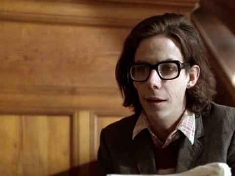 A young man with long dark hair and thick rimmed glasses