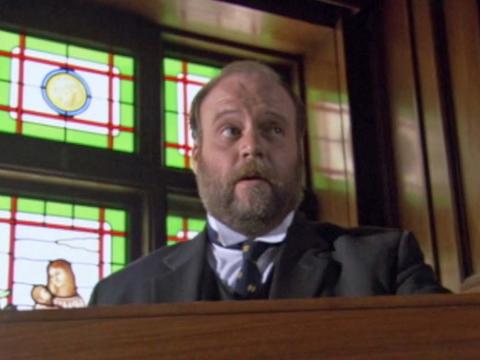 A man in a suit and tie speaks at a podium with a stained glass window behind him