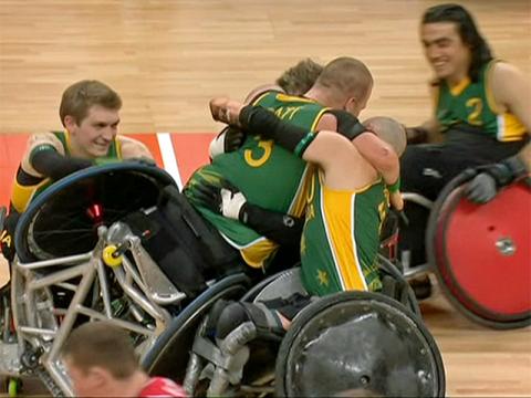 Three wheelchair rugby players embrace on an indoor rugby court at the Paralympics. They are all wearing green and gold uniforms.