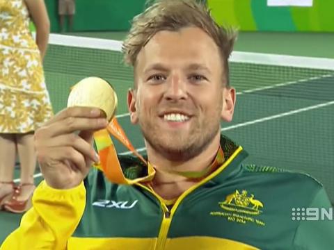 Wheelchair tennis player Dylan Alcott smiles for the camera. He is holding a gold medal and smiling. He is wearing an Australian green and gold uniform.