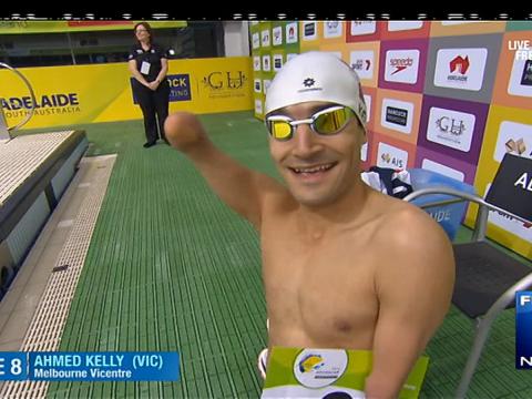 Paralympic swimmer Ahmed Kelly is waving at the camera. His arm is amputated below his elbow on both of his arms. He is wearing goggles and a swimming cap.