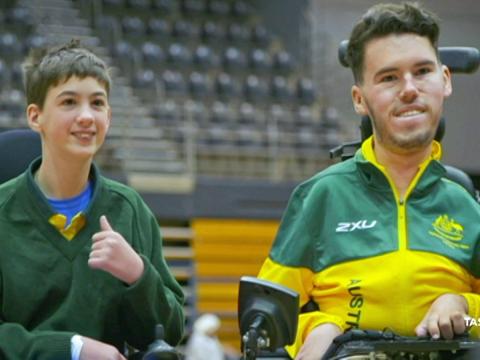 Boccia player Dan Michel is posing for a photo in his wheelchair next to a child who is also in a wheelchair. Michel wears a green and gold uniform.