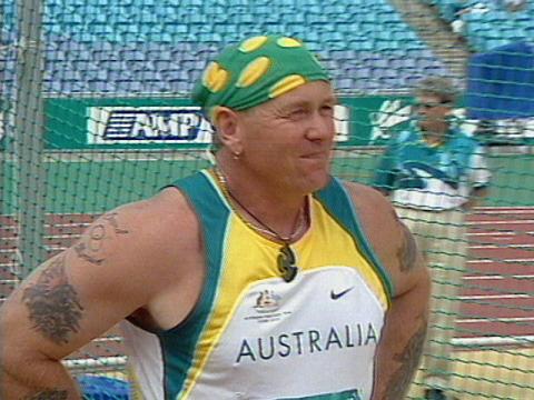 Paraathlete John Eden is wearing green and gold. His arms are tattooed and he looks out over a field at Olympic stadium. 