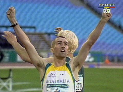 Paraathlete Tim Sullivan is wearing green and gold. He is holding his arms up in the air at an Olympic stadium.