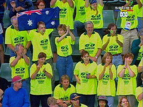 A number of sports fans wear green and gold t-shirts and stand cheering in the stands at a sporting event. One man is holding an Australian flag up above his head.