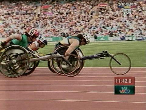 Wheelchair racer Louise Sauvage is powering ahead of a pack of other paraathletes on a racing track. There are thousands of spectators watching on.