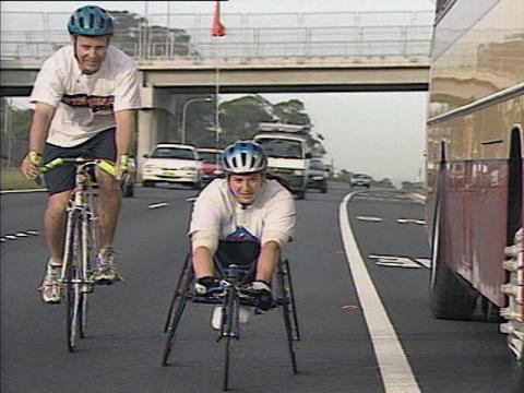 Wheelchair racer Louise Sauvage is riding on the road with a cyclist next to her. There are cars and a bus nearby but she has plenty of room on the road.