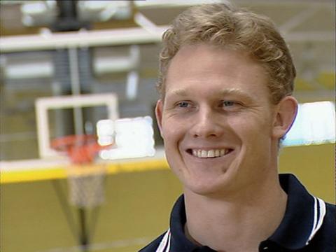 Wheelchair basketballer Troy Sachs is smiling for the camera in an interview setting. There is a basketball hoop in the background of the shot.