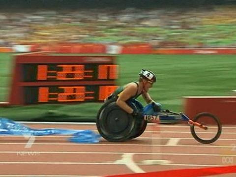 Wheelchair racer Kurt Fearnley is captured having just crossed the finish line in the marathon at the Beijing 2008 Paralympics. A blurred time board and a packed stadium are blurred behind him.