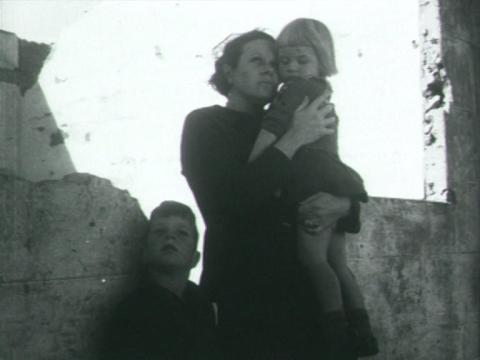A woman standing in a ruined building holds her children