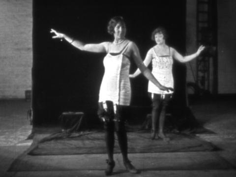 Two women wearing corsets pose with their arms up in the air. They are in a studio with a black backdrop.