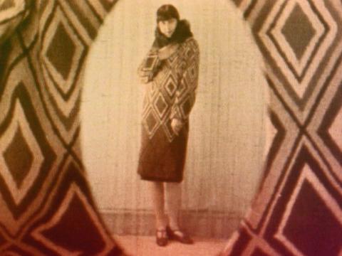 An orange tinted black and white still of a woman wearing a diamond-patterned dress with a fur cuff around her neck. The shot is framed by an oval cut out of matching diamond patterned material.