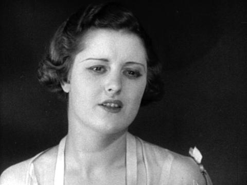 Black and white still frame of a woman with very thin eyebrows speaking. She has a 1930s hairstyle in keeping with the period.