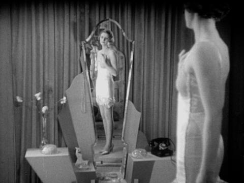 A women wearing a white Berlei foundation garment is looking at her reflection in an art deco mirror.