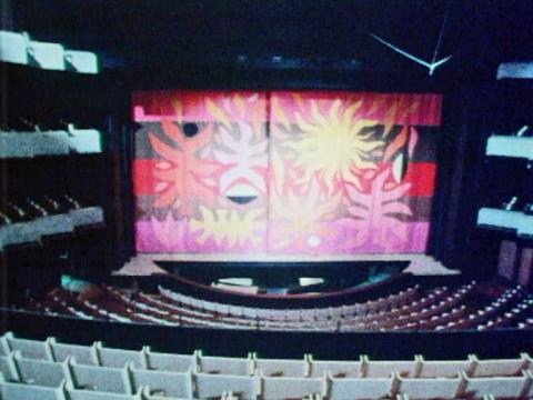 Inside the concert hall of the Sydney Opera House with empty chairs all around. There is a colourful backdrop on the stage.