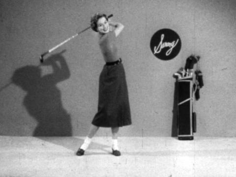 A woman swings a golf club cheerfully while smiling at the camera. She is in a studio setting.
