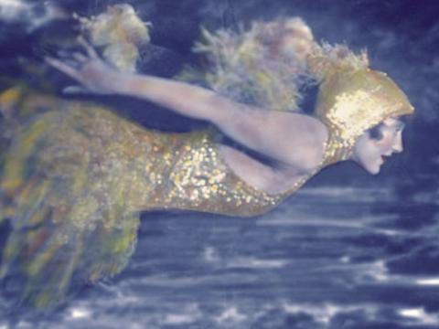 Hand-tinted photograph of Annette Kellerman swimming underwater wearing an elaborate costume including ballet slippers and head-dress.