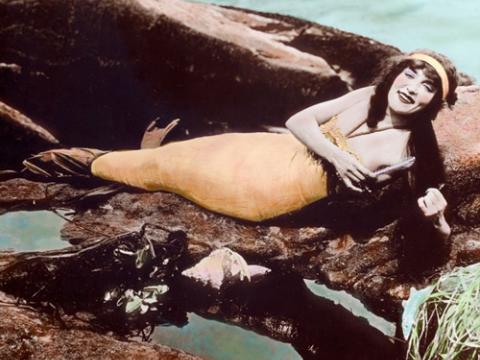 A hand-tinted photograph of Annette Kellerman wearing a mermaid costume and lying on rocks near the water.