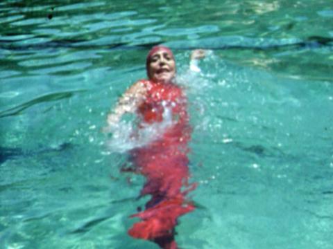 Annette Kellerman as an older woman wearing a bathing suit and swimming cap. She is swimming on her back in a pool and looking at the camera.