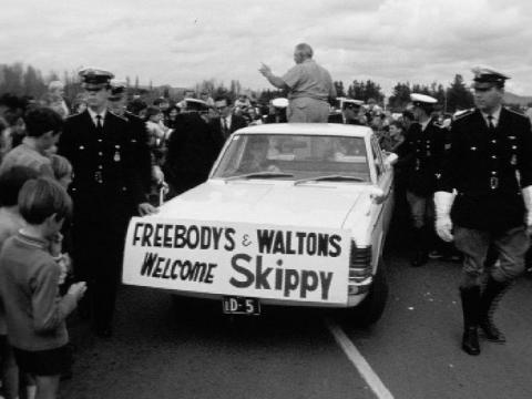 A car moves though a crowd with a banner reading, 'Freebody's and Waltons welcome Skippy'.