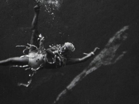 Annette Kellerman is performing an underwater ballet, dressed in a swimming costume adorned with baubles. She is striking a dramatic pose with her arms and legs outstretched.