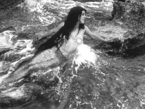 Annette Kellerman is in a mermaid costume, posing on a rock as the ocean's waves crash against her.