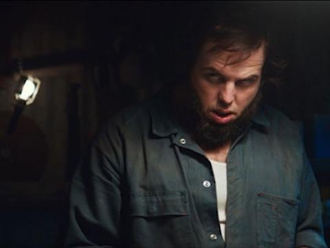 A man in overalls in a dark room with a malcious expression.