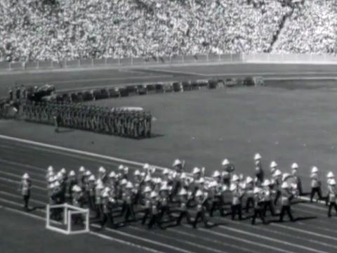 Marching band and rows of athletes on a field inside a stadium full of people during the opening ceremony of the 1962 Commonwealth Games in Brisbane