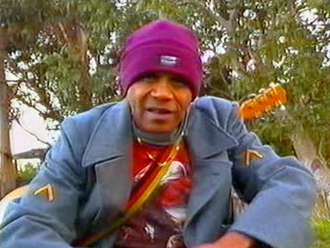 Musician and songwriter Archie Roach wearing a blue coat and purple beanie, pictured from the waist up, carrying a guitar on his back and looking directly down into camera.
