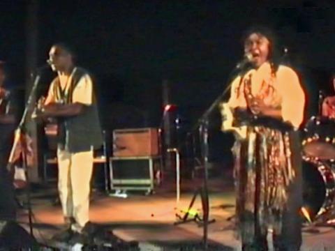 Indigenous musicians Archie Roach and Ruby Hunter pictured on stage performing. Archie is holding a guitar and Ruby is holding a tambourine and both are singing into microphones.